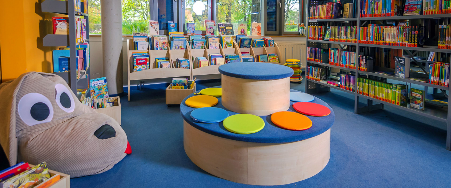 Aufnahme der Kinderbibliothek mit rundem Sitzpodest, Bücherregalen und einem Sitzsack in Form eines Hundegesichts.
