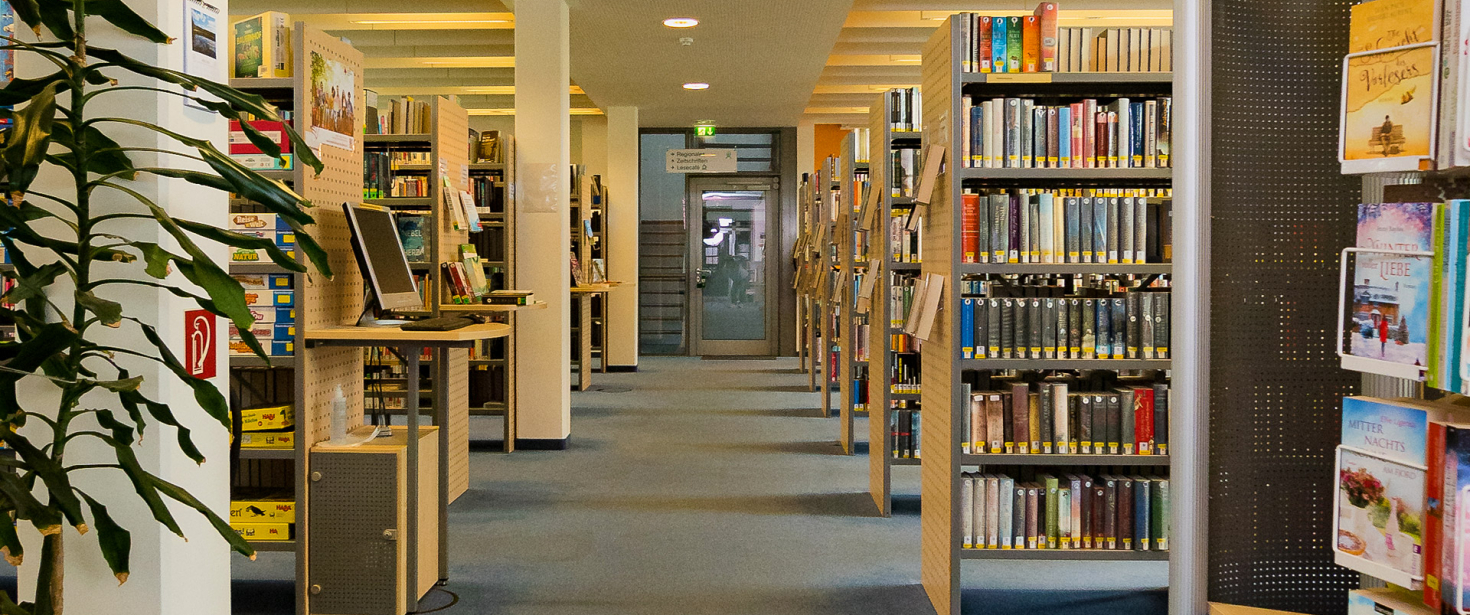 Aufnahme des Mittelgangs der Bibliothek. Links und rechts Regale sowie ein Recherche-PC.