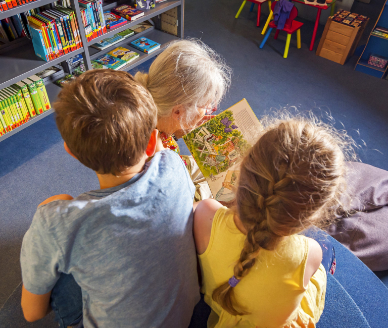 Eine Frau und zwei Kinder lesen gemeinsam ein Buch auf dem Podest in der Kinderabteilung.